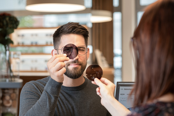 Man with a beard tests round sunglasses at a store counter, while a salesperson holds up another pair to compare.