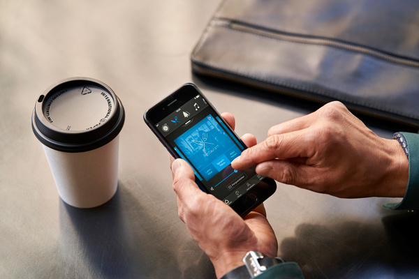 Hands hold a smartphone displaying a blue map/blueprint on the screen beside a takeaway coffee cup on a metal table.
