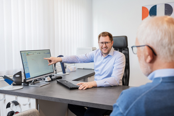Office scene: man in a light blue shirt points at a computer screen while consulting with an older man wearing glasses at the desk