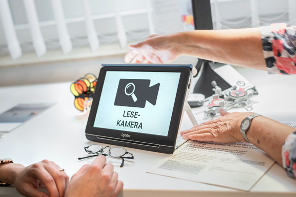 Assistive reading camera device on a white desk, displaying a magnifying-camera icon and the words Lese-Kamera, with hands nearby and reading glasses nearby.