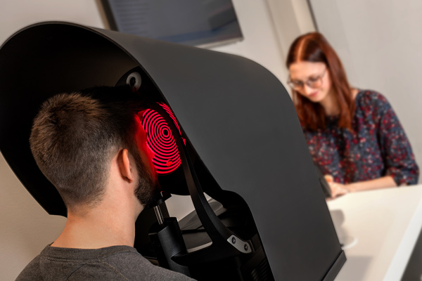 Person sits inside a large black optical imaging device with red circular light, while a technician watches from a desk nearby.